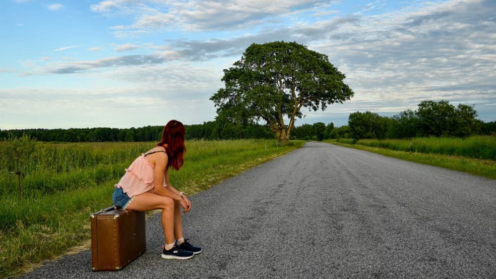 A woman in denim shorts and pink top sits on luggage waiting by the road in Estonia.
